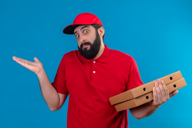 Confuso giovane uomo di consegna caucasico bello che indossa l'uniforme rossa e cappuccio che tiene le scatole per pizza e mostrando la mano vuota isolata sull'azzurro