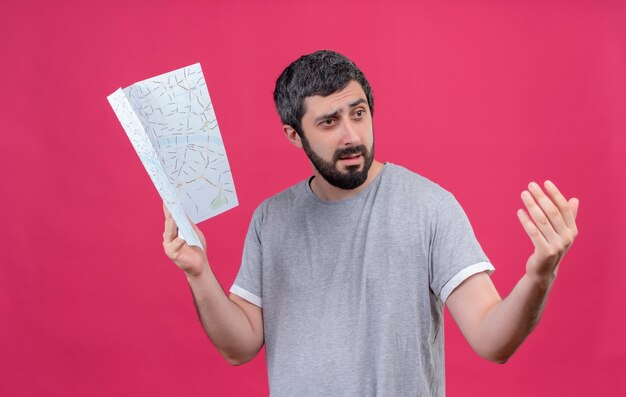 Confused young handsome caucasian traveler man holding map looking at side and doing come here gesture isolated on pink 