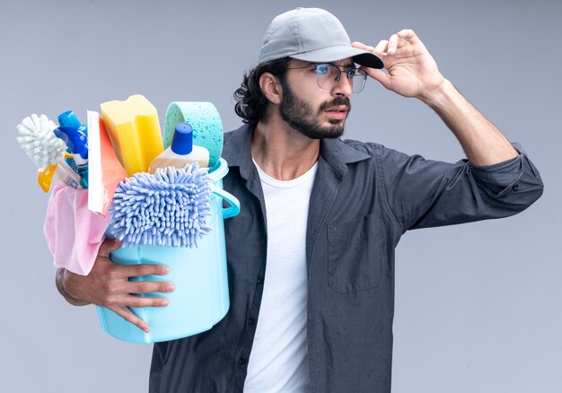 Confused young handsome cleaning guy wearing t-shirt and cap holding bucket of cleaning tools and holding cap isolated on white wall