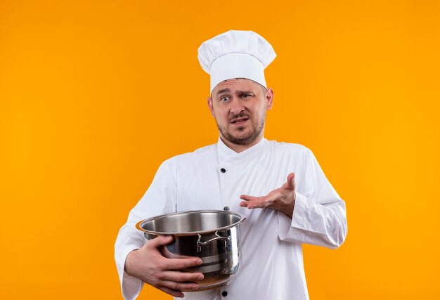 Смущенный молодой красивый повар в униформе шеф-повара держит котел и показывает пустую руку на изолированной оранжевой стене