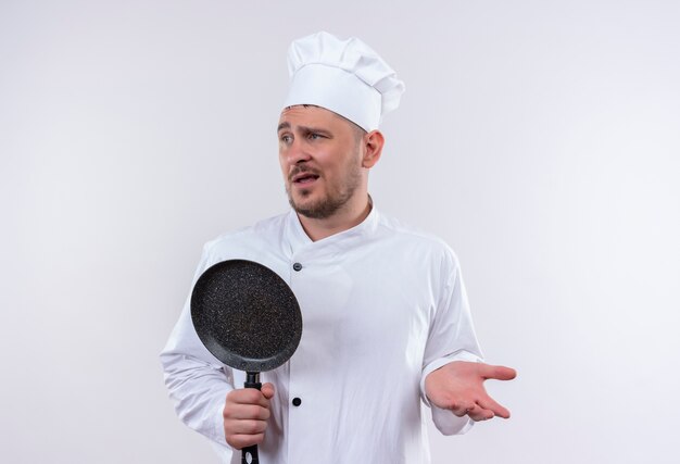 Confused young handsome cook in chef uniform holding frying pan showing empty hand and looking at side isolated on white wall