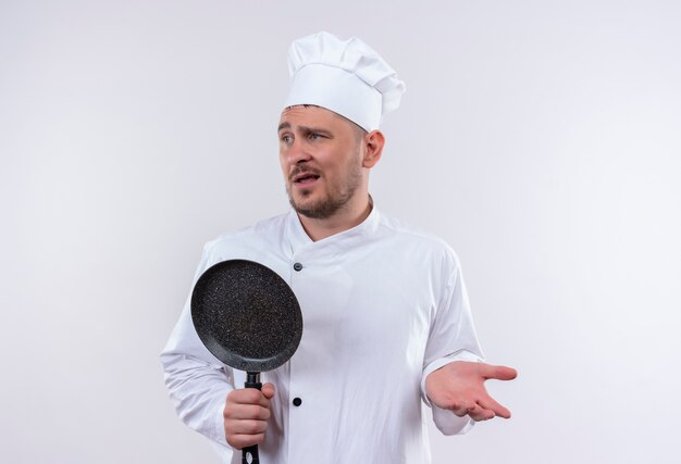 Смущенный молодой красивый повар в униформе шеф-повара держит сковороду, показывая пустую руку и смотрит в сторону, изолированную на белой стене