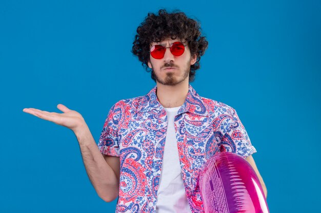 Confused young handsome curly man wearing sunglasses holding swim ring showing empty hand on isolated blue space