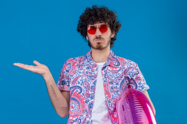 Confused young handsome curly man wearing sunglasses holding swim ring showing empty hand on isolated blue space