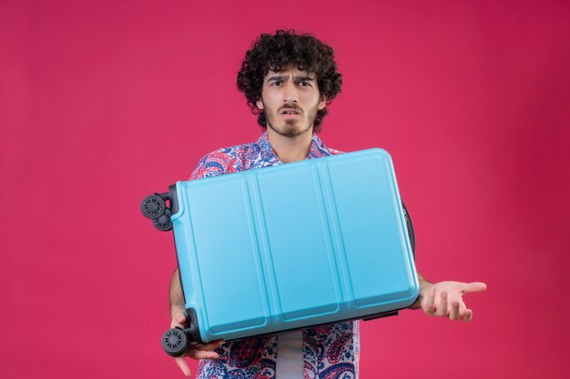 スーツケースを持って、コピースペースと孤立したピンクのスペースに空の手を示す混乱した若いハンサムな巻き毛の旅行者