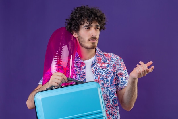 Смущенный молодой красивый кудрявый путешественник держит чемодан и кольцо для плавания, указывая на правую сторону на изолированном фиолетовом пространстве