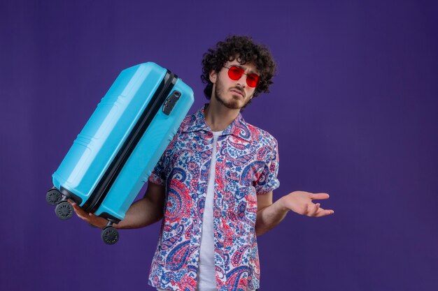コピースペースと孤立した紫色のスペースに空の手を示すスーツケースを保持しているサングラスを身に着けている混乱した若いハンサムな巻き毛の旅行者の男