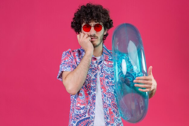 Confused young handsome curly traveler man wearing sunglasses holding swim ring with hand on chin on isolated purple space with copy space