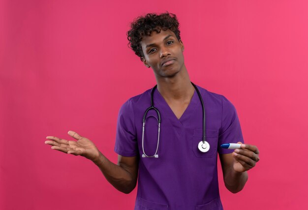 A confused young handsome dark-skinned doctor with curly hair wearing violet uniform with stethoscope holding thermometer 