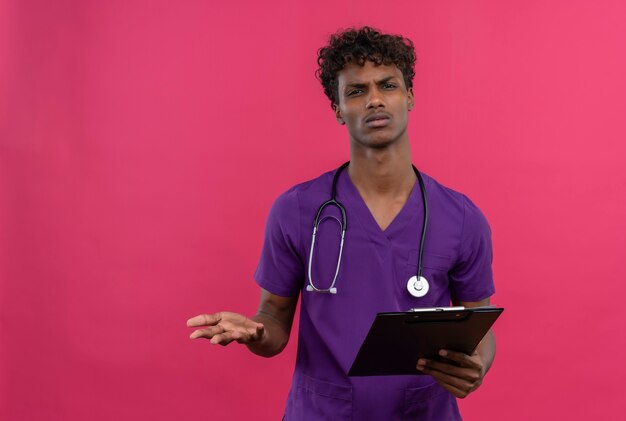 A confused young handsome dark-skinned doctor with curly hair wearing violet uniform with stethoscope looking angry at camera while holding clipboard 