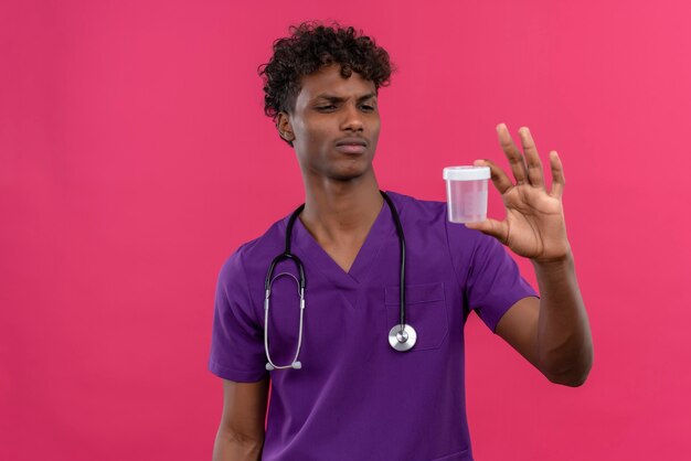 A confused young handsome dark-skinned doctor with curly hair wearing violet uniform with stethoscope looking at medical plastic specimen jar 
