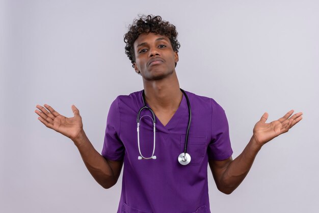 Смущенный молодой красивый темнокожий доктор с кудрявыми волосами в фиолетовой форме со стетоскопом раскрывает руки и не знает, что делать.