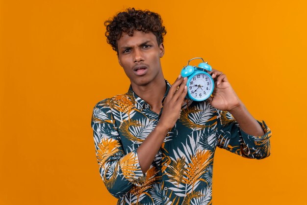 Confused young handsome dark-skinned man with curly hair in leaves printed shirt holding blue alarm clock and showing time 