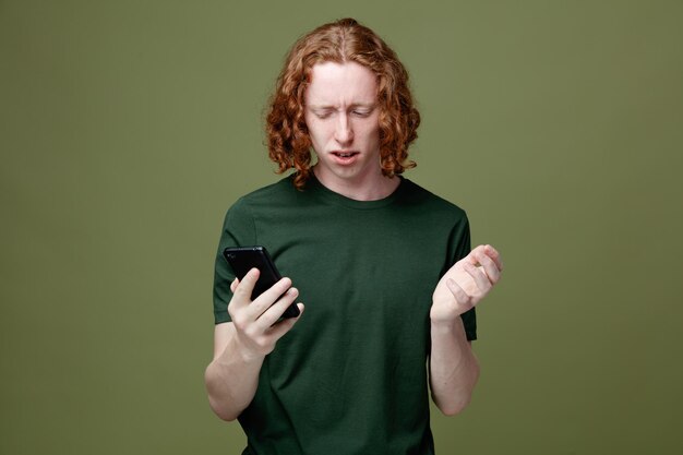 緑の背景に分離された緑の t シャツを着て携帯電話を見て混乱している若いハンサムな男