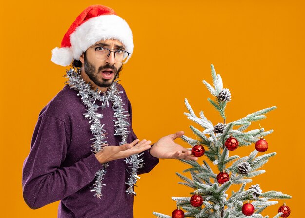 Confused young handsome guy standing nearby christmas tree wearing christmas hat with garland on neck holding and points at tree isolated on orange wall