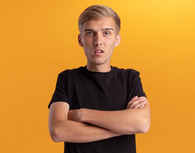 Confused young handsome guy wearing black shirt crossing hands isolated on yellow wall