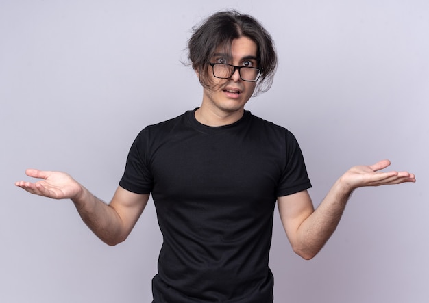 Confused young handsome guy wearing black t-shirt and glasses spreading hands isolated on white wall
