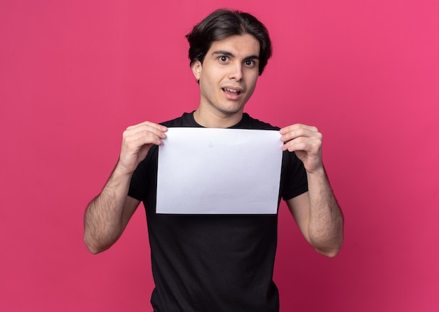 Free photo confused young handsome guy wearing black t-shirt holding paper isolated on pink wall