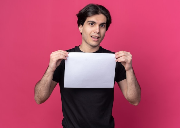 Confused young handsome guy wearing black t-shirt holding paper isolated on pink wall