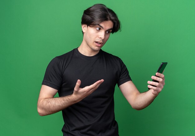 Confused young handsome guy wearing black t-shirt holding and points with hand at phone isolated on green wall