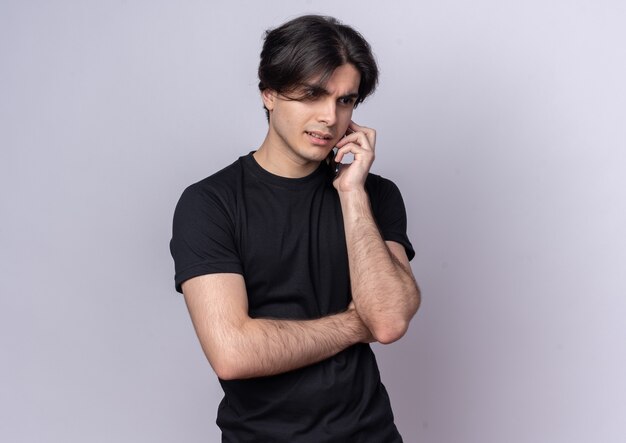 Смущенный молодой красивый парень в черной футболке говорит по телефону, изолированному на белой стене