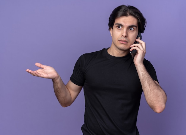 黒のTシャツを着て混乱している若いハンサムな男は電話で話し、紫色の壁に隔離された手を広げます