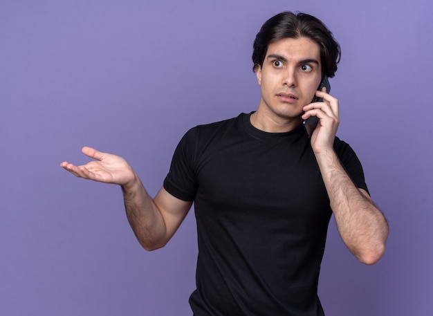 Confused young handsome guy wearing black t-shirt speaks on phone and spreading hand isolated on purple wall