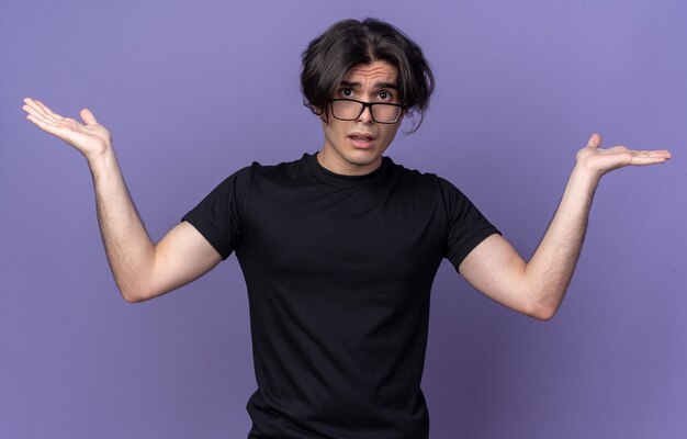 Confused young handsome guy wearing black t-shirt spreading hands isolated on purple wall