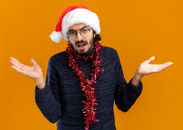 Confused young handsome guy wearing christmas hat with garland on neck spreading hands isolated on orange background