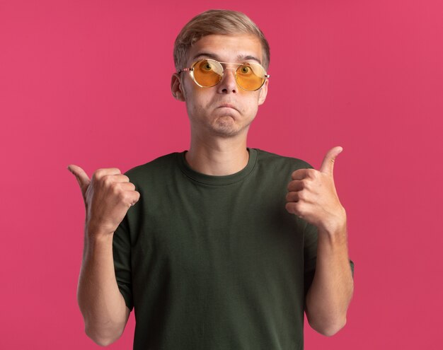Confused young handsome guy wearing green shirt and glasses points at differents sides isolated on pink wall with copy space