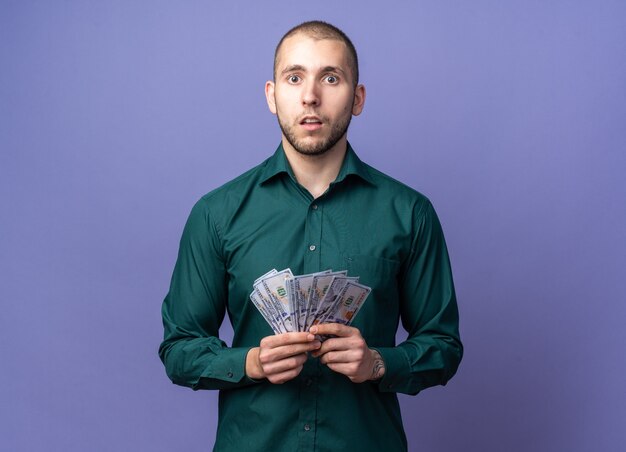 オリーブグリーンの壁に隔離された正面を見てお金と医療薬のパックを保持しているマスクを身に着けている混乱した若いハンサムな金髪の病気の男 無料の写真