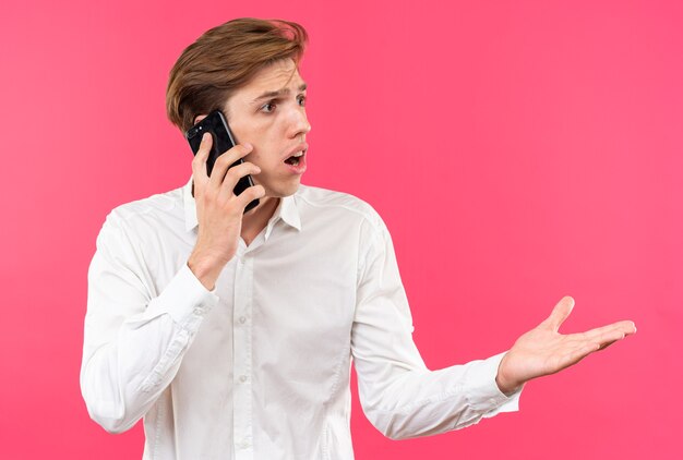 Confused young handsome guy wearing white shirt speaks on phone points with hand at side 