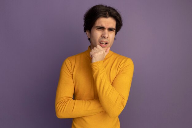 Смущенный молодой красивый парень в желтом свитере с высоким воротом, положив руку под подбородок, изолирован на фиолетовой стене