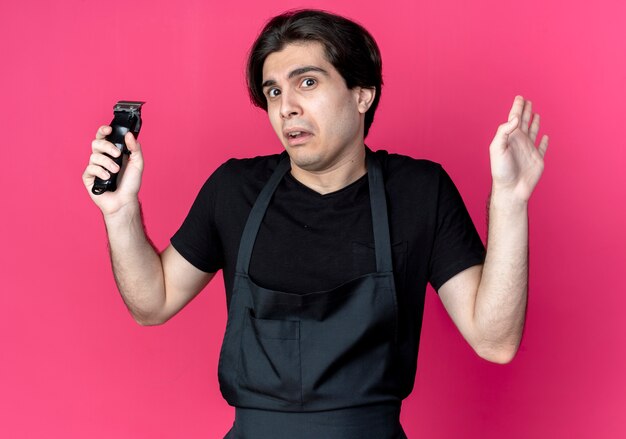 Confused young handsome male barber in uniform holding hair clippers and spread hand 