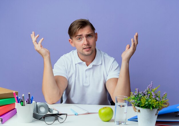 confused young handsome male student sitting at desk with school tools spreads hands