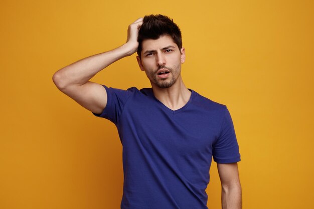 Confused young handsome man keeping hand on head looking at camera on yellow background