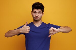 Confused young handsome man looking at camera showing thumbs up and down on yellow background