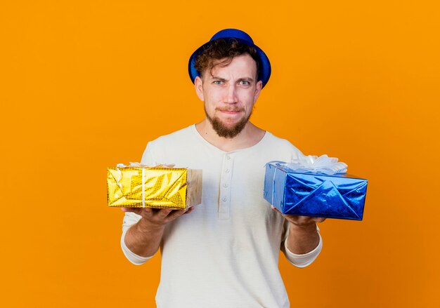 Confused young handsome slavic party guy wearing party hat holding gift boxes looking at camera isolated on orange background with copy space