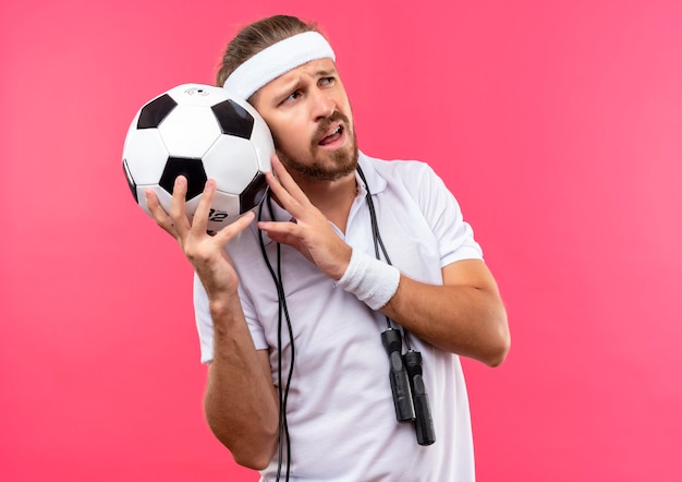 Confused young handsome sporty man wearing headband and wristbands holding soccer ball looking at side with jump rope around neck isolated on pink wall