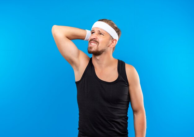 Confused young handsome sporty man wearing headband and wristbands putting hand behind head looking up isolated on blue wall