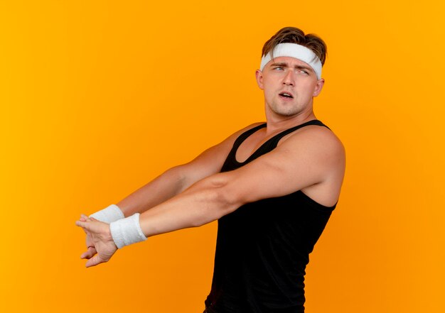 Confused young handsome sporty man wearing headband and wristbands standing in profile view looking at side and stretching hands isolated on orange 