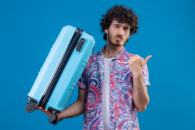 Confused young handsome traveler man holding suitcase pointing at right side on isolated blue space with copy space