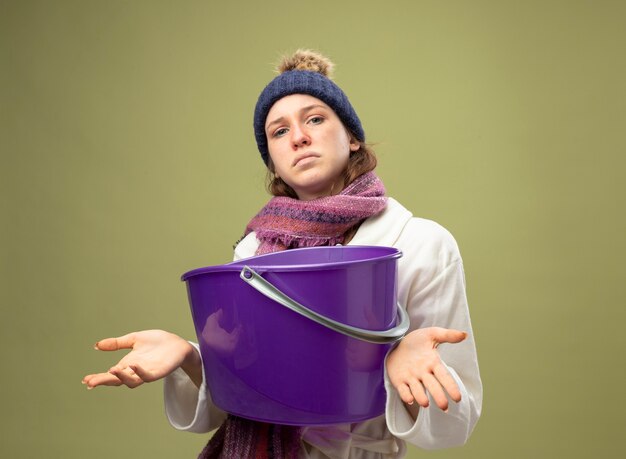 Confused young ill girl wearing white robe and winter hat with scarf holding plastic bucket spreading hands isolated on olive green