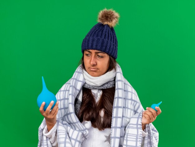 Confused young ill girl wearing winter hat with scarf holding and looking at enemas isolated on green background