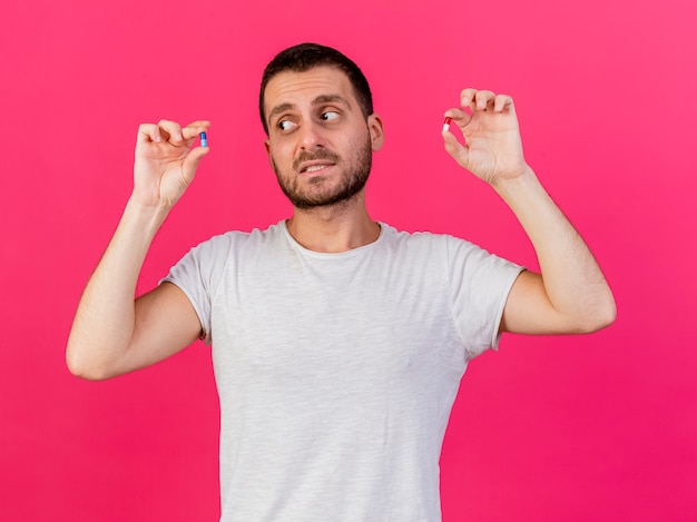 Confused young ill man raising and looking at pills isolated on pink background