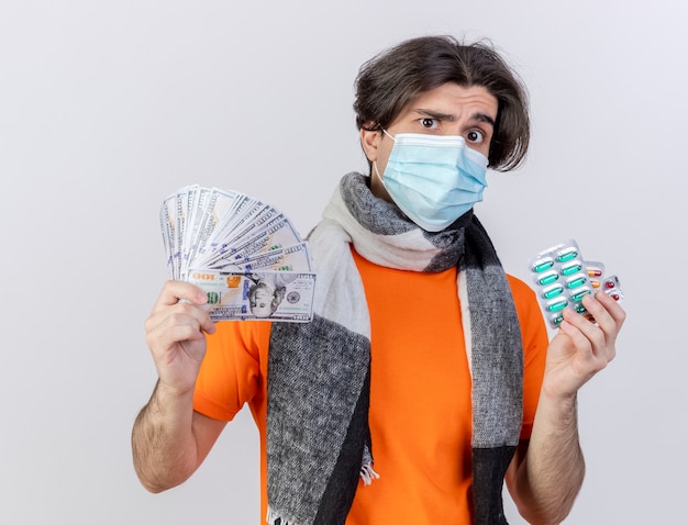 白い背景で隔離の丸薬と現金を保持しているスカーフと医療マスクを身に着けている混乱した若い病気の男