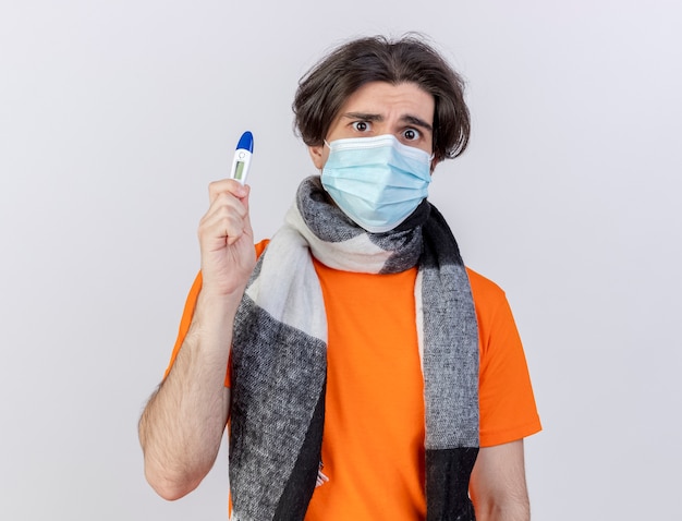 Confused young ill man wearing scarf and medical mask holding thermometer isolated on white background