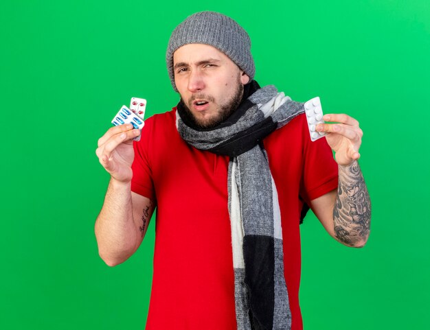 Confused young ill man wearing winter hat and scarf holds packs of medical pills isolated on green wall