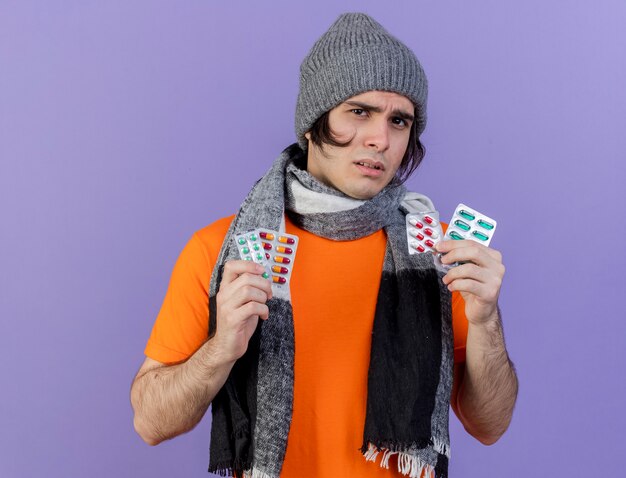 Confused young ill man wearing winter hat with scarf holding pills isolated on purple background