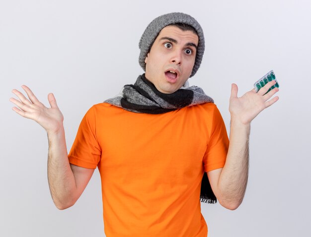 Смущенный молодой больной человек в зимней шапке с шарфом держит таблетки и разводит руками, изолированными на белом фоне